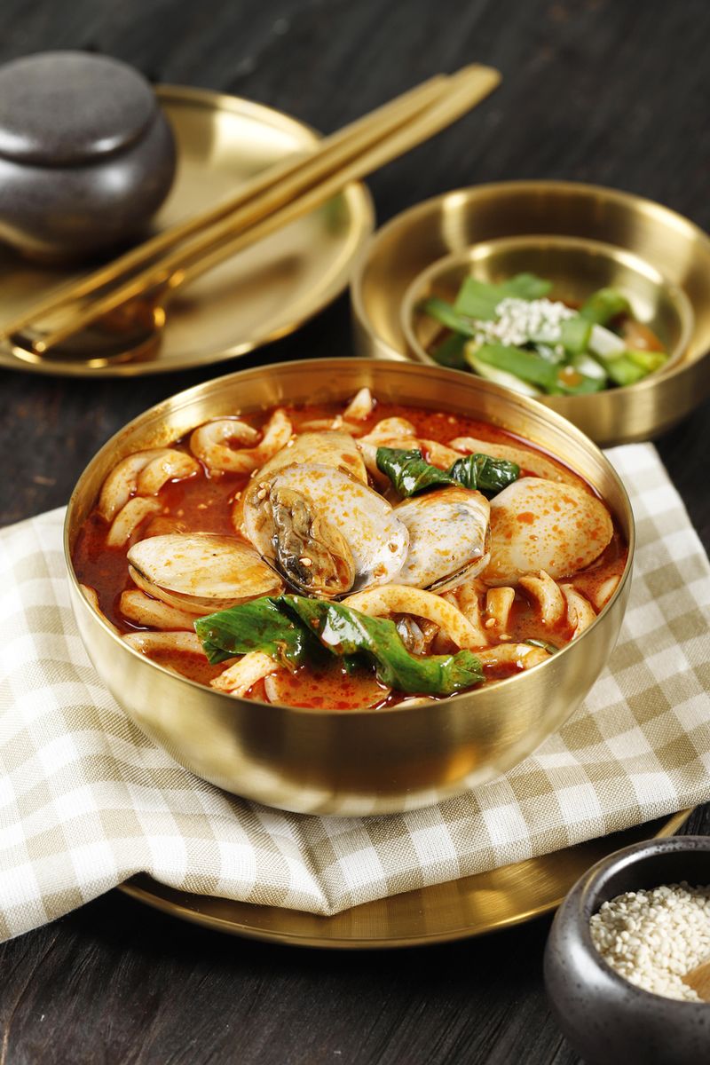 Red Seafood Jjampong, Red Spicy Seafood Korean Noodle Soup with Vegetable and Clam on Golden Bowl, Above Wooden Table