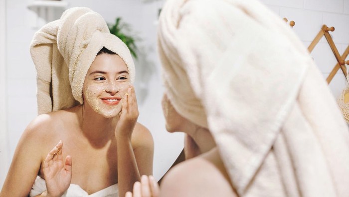 Skin Care concept. Young happy woman in towel making facial massage with organic face scrub and looking at mirror in stylish bathroom. Girl applying scrub cream, peeling and cleaning skin