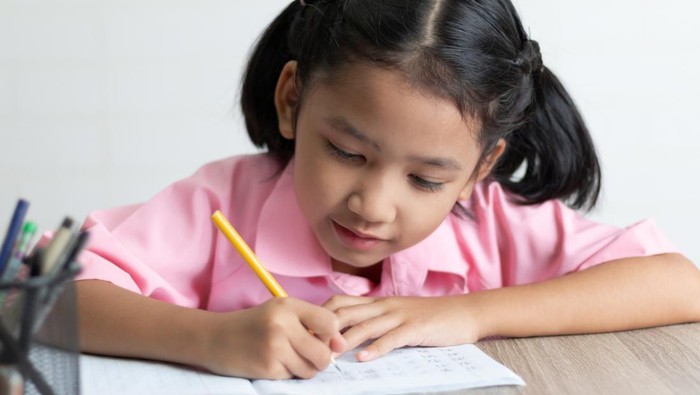 Close up the little girl is doing homework happily. Children use a yellow pencil is writing a notebook on the wooden table. Select focus shallow depth of field.