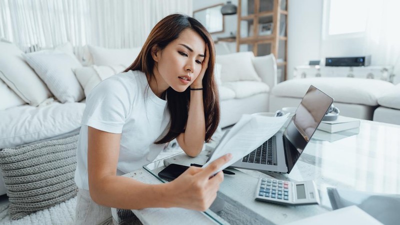Young Asian woman handling home finances with laptop, looking worried while going through financial bills. Financial plan, tax, spending and budgets, financial problems concept
