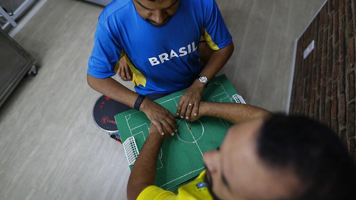Helio Fonseca, right, helps his deaf and blind friend Carlos Junior, center, with a play-by-play of the World Cup group G soccer match between Brazil and Switzerland, in Osasco, Sao Paulo state, Brazil, Monday, Nov. 28, 2022. Fonseca uses a tactile table and touch on the body of visually and hearing-impaired people to show them the movements of the ball and of the players during soccer matches. Brazil won the match 1-0. (AP Photo/Marcelo Chello)
