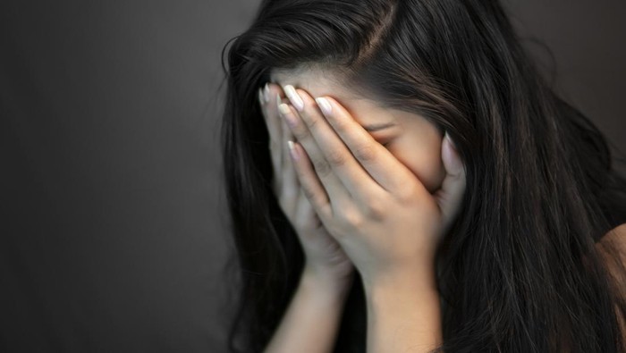Close-up portrait of alone, stressed young woman sitting in darkness and crying with covering her face with hands.