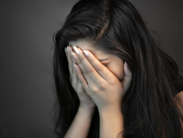 Close-up portrait of alone, stressed young woman sitting in darkness and crying with covering her face with hands.