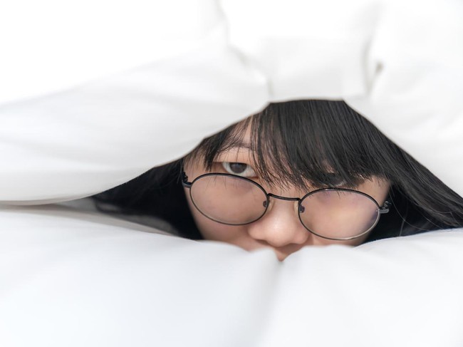 close up to the Cute Asian Glasses woman, she hides her body inside white pillow on the bed