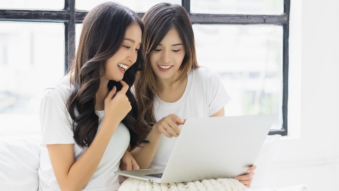 Two beautiful asian women using laptop computer talking and smiling while sitting on bed at home Happy girls using laptop for Internet and social media Beautiful young lady enjoy using laptop computer