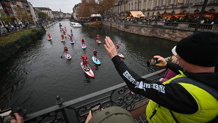 Sambut Natal, Rombongan Sintreklas Dayung Papan di Sungai Prancis Pestrians take picture of paddlers dressed as Santa Claus as they attend a SUP (Stand Up Paddleboarding) parade on the Ill river in Strasbourg, eastern France, on December 3, 2022. (Photo by SEBASTIEN BOZON / AFP) (Photo by SEBASTIEN BOZON/AFP via Getty Images)
