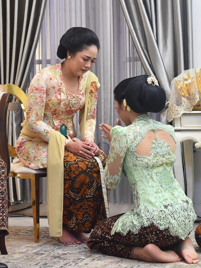 Erina sudah menanti kehadiran mereka dalam balutan kebaya kutubaru berwarna hijau kebiruan penuh detail brokat rancangan Intan Avantie yang juga putri desainer kondang Anne Avantie. Hijau itu lambang harapan dan kesuburan. Mudah-mudahan kedua mempelai harapannya dikabulkan dan mempunyai keturunan, ujar Wigung saat memandu prosesi tersebut. Kebaya tersebut dilengkapi stagen jumputan merah dan bawahan berupa jarik batik motif sidoluhur. Penampilan Puteri Indonesia Yogyakarta 2022 itu kian ayu dengan sanggul. Foto: Tim Media Pernikahan Erina-Kaesang/Kris