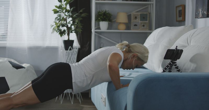 Medium shot of a woman doing push-ups of a sofa at home while looking at smartphone