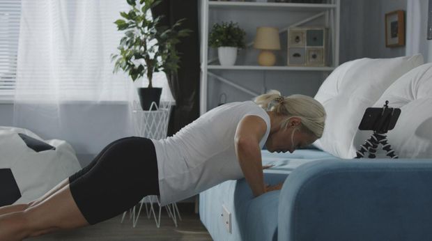 Medium shot of a woman doing push-ups of a sofa at home while looking at smartphone