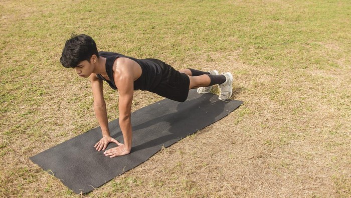A young man does a set of diamond pushups on a black mat at an open field outdoors. Chest or full body workout.