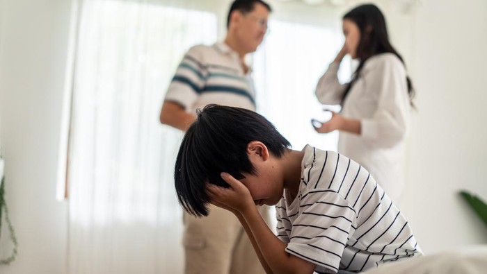 Ilustrasi Dampak Anak Broken Home Asian boy kid sitting and crying on bed while parents having fighting or quarrel conflict at home. Child covering face and eyes with hands do not want to see the violence. Domestic problem in family.