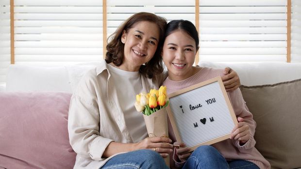 Attractive beautiful asian middle age mum sit with grown up daughter give gift box and flower in family moment celebrate mother day. Overjoy bonding cheerful kid embrace relationship with retired mom.