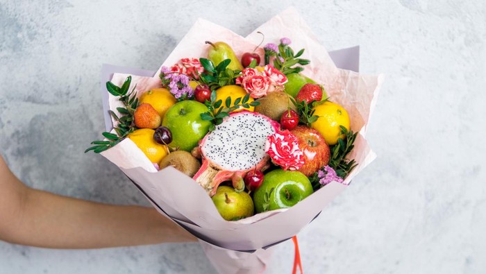 Fruit and berries bouquet. Eating bouquet in female hand on gray background. Apple, orange, strawberry, pear, kiwi, dragon fruit and flowers, eucalyptus. Shallow DOF. Copy space