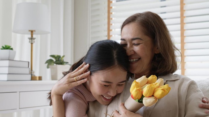 Attractive beautiful asian middle age mum sit with grown up daughter give gift box and flower in family moment celebrate mother day. Overjoy bonding cheerful kid embrace relationship with retired mom.