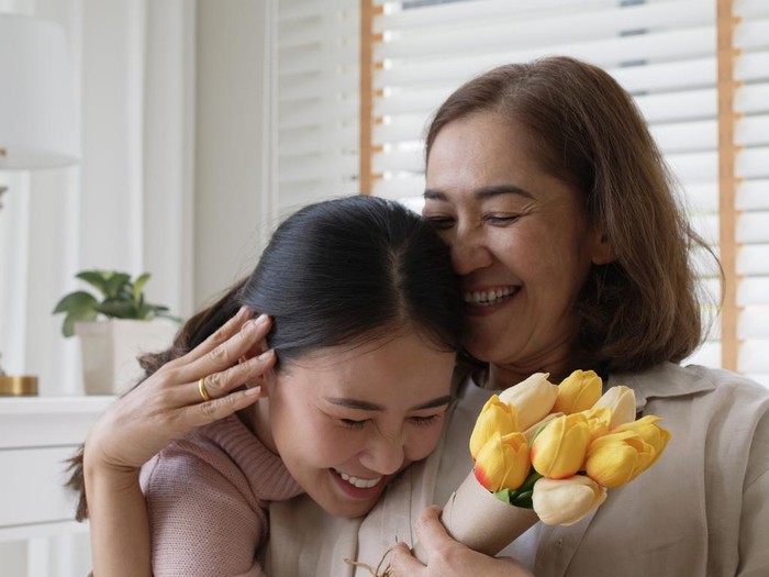 Attractive beautiful asian middle age mum sit with grown up daughter give gift box and flower in family moment celebrate mother day. Overjoy bonding cheerful kid embrace relationship with retired mom.