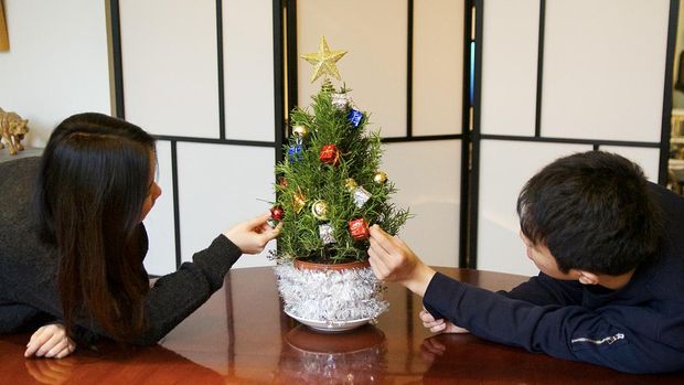 Youthful Asian students decorate a miniature Christmas tree with gold star, bells and colorful packages on table in front of checkered blind