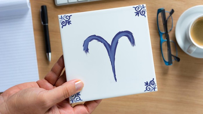 Person holding Delfts Blue aries zodiac tile above working desk. Drawing on white tile by contributor