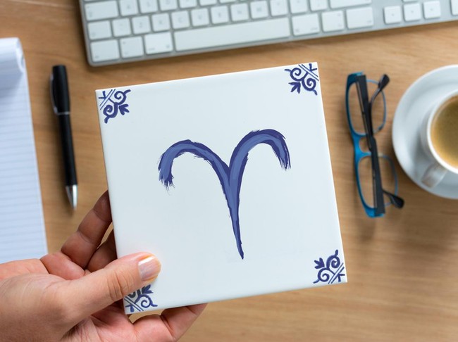 Person holding Delfts Blue aries zodiac tile above working desk. Drawing on white tile by contributor