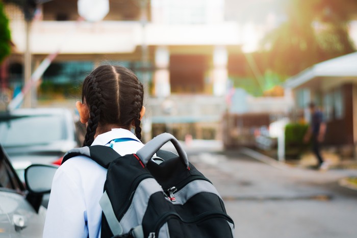 Little girl walking into school with backpack in the morning under warm sunshine. Education concept.