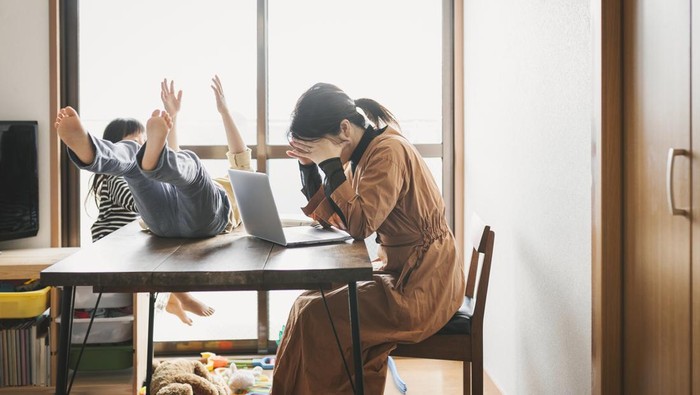 Asian mother working with laptop while her son and daughter are playing in the living room. Her son is playing on the table.