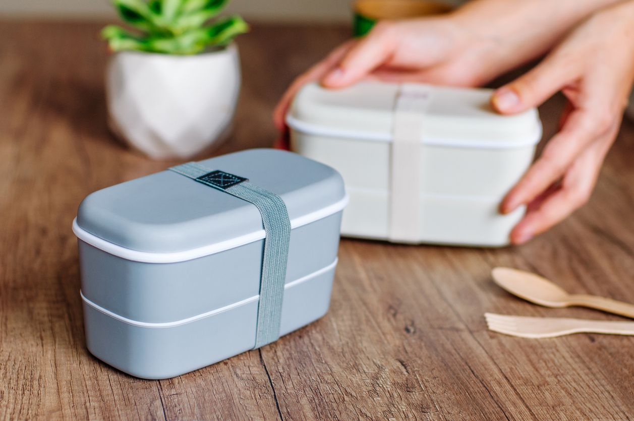 Two closed plastic two-layered lunch boxes with elastic band near wooden forks and spoons and paper cup on wooden table. Female hands holding one lunch box on the background. Selective focus on lunch box on the table.