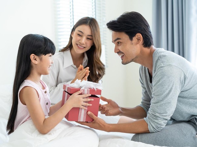 Asian lovely young little daughter open present gift box from parents. Happy family, kid feel happy and excited for surprise from mother and father ready to celebrate Birthday party together at home.