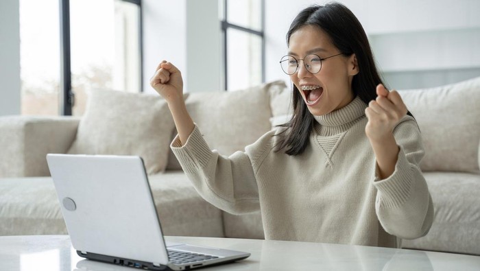 A successful Asian businesswoman with glasses enjoys success with a laptop on her desktop. A joyful woman who has received a jackpot celebrating her victory