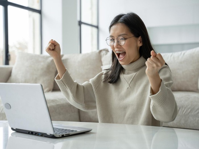 A successful Asian businesswoman with glasses enjoys success with a laptop on her desktop. A joyful woman who has received a jackpot celebrating her victory