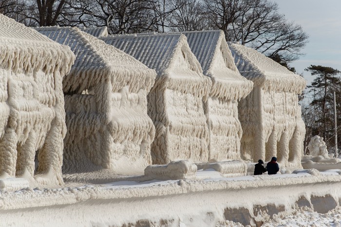 People walk by homes covered in ice at the waterfront community of Crystal Beach in Fort Erie, Ontario, Canada, on December 28, 2022, following a massive snow storm that knocked out power in the area to thousands of residents. (Photo by Cole Burston / AFP) (Photo by COLE BURSTON/AFP via Getty Images)