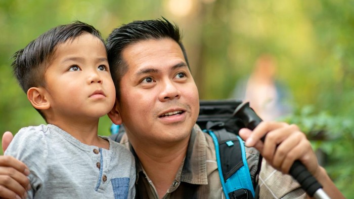 A Filipino father and son take a hike in the woods birdwatching.  The father has crouched down low to the young boy as they both look to the trees at the birds.