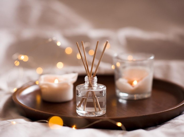 Home perfume in glass bottle with wood sticks, scented burn candles  tray in bedroom close up. Aromatherapy cozy atmosphere lifestyle. Winter warm xmas season.