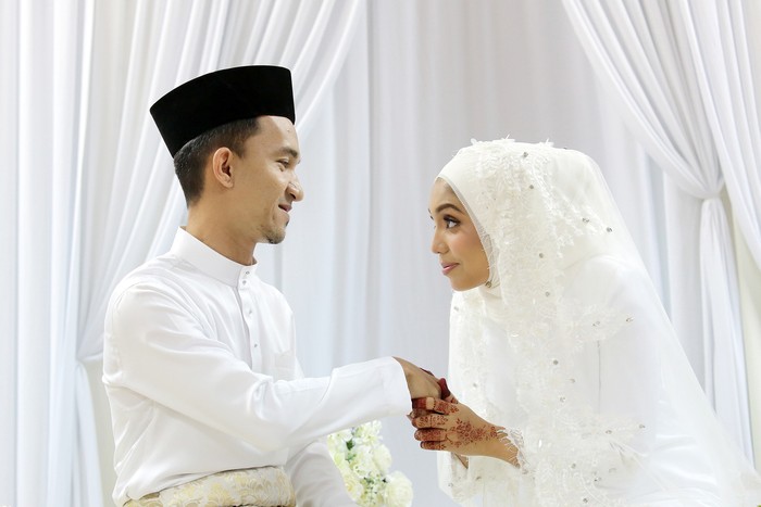 A Muslim young woman holding a young mans hand for salam (gesture of mannerism in greeting) when they are certified newlywed in Islam akad nikah (solemnisation) ceremony. In this ceremony, newlywed and all guests are required to wear pure white tradiitonal clothing.