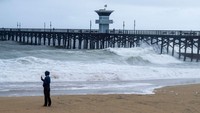 Seorang pria mengambil foto saat ombak menerjang bawah Dermaga Pantai Seal tatkala badai musim dingin membawa angin, hujan, dan banjir ke Orange County. Mark Rightmire/MNG/Getty Images