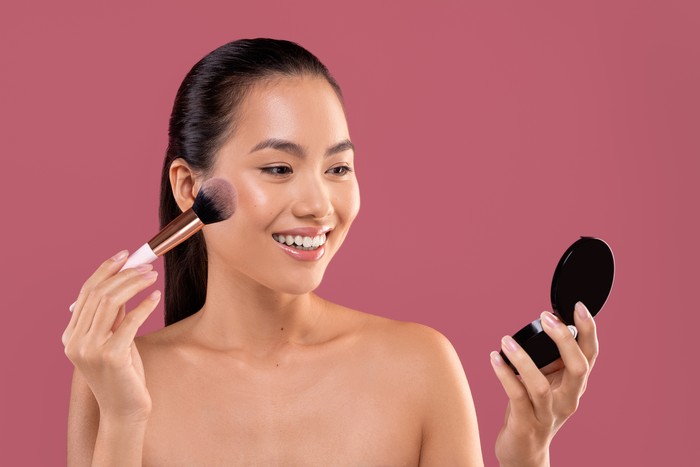 1374583158 Pretty topless young chinese woman with makeup brush looking at mirror, applying blush on her cheeks, enjoying brand new makeup products on purple studio background, closeup photo