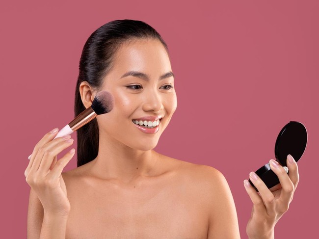 Pretty topless young chinese woman with makeup brush looking at mirror, applying blush on her cheeks, enjoying brand new makeup products on purple studio background, closeup photo