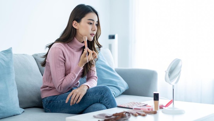 Pretty topless young chinese woman with makeup brush looking at mirror, applying blush on her cheeks, enjoying brand new makeup products on purple studio background, closeup photo