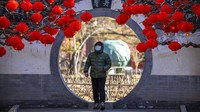 Seorang pria yang mengenakan masker berjalan melewati pepohonan yang dihiasi lentera untuk Tahun Baru Imlek di sebuah taman umum di Beijing, China, Jumat(20/1/2023).