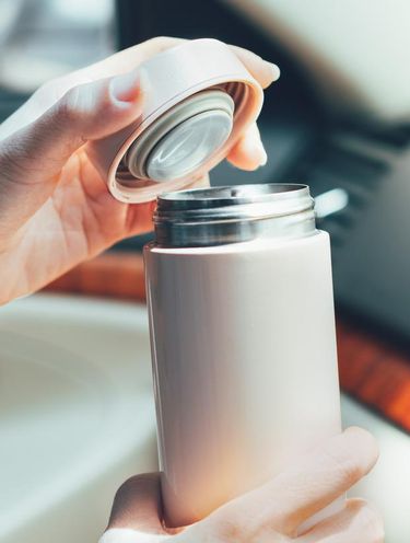 1392673482 Close up of woman's hands holding reusable insulated water bottle and opening the lid in car in bright sunlight