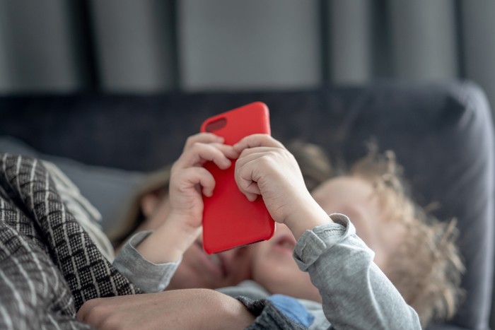 Mom and toddler son playing with smartphone, lying on the sofa under blanket. Screen time limit, enjoying time together at home.
