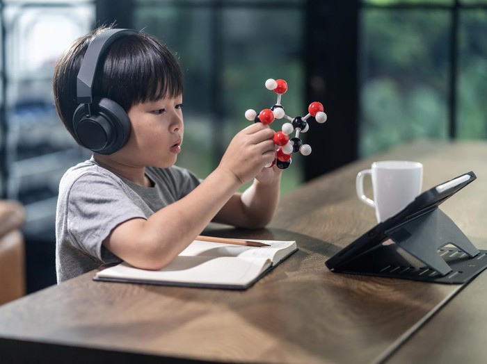 Asian little boy assembling molecular model by learning from home with school teacher teaching online. Homeschool kid practicing to think about logic and conference with teacher by remote application.