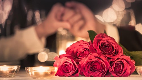 Couple at a candle light dinner date holding hands next to bouquet of red roses.