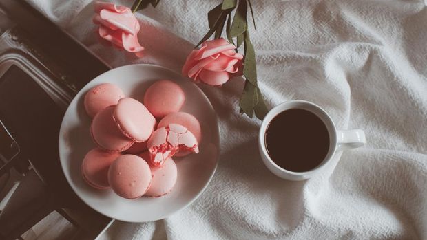 Pink macaron cookies with cup of coffee and rose flowers on white background. Spring still life scene. Vintage pastel styled photo. Copy space for you text. Toned image.