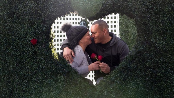Sepasang kekasih berfoto di belakang instalasi cinta sembari memegang bunga mawar.