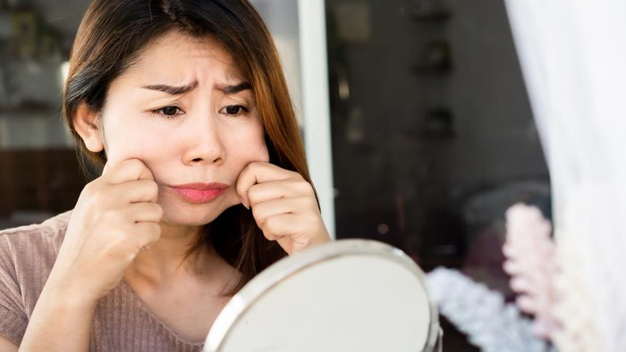 worried Asian woman hand pulling her fat skin on cheeks ,checking her face on mirror ,aging and overweight concept