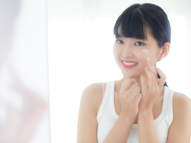 Beautiful young asian woman looking in mirror and apply moisturizer with cosmetic cream or lotion in bathroom at home, skincare for hygiene, treatment and bodycare, skin and anti-aging of facial.