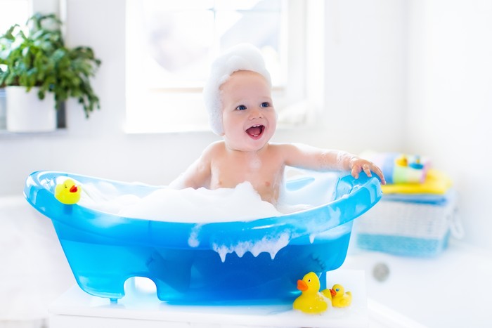 Happy laughing baby taking a bath playing with foam bubbles. Little child in a bathtub. Smiling kid in bathroom with colorful toy duck. Infant washing and bathing. Hygiene and care for young children.