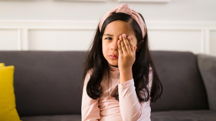 Portrait of a sick little child suffering and in pain covering her eye with a hand because of an eye infection