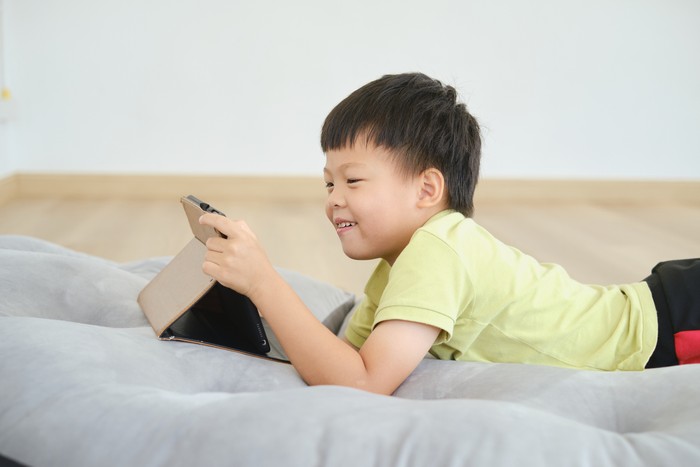 Cute little Asian kindergarten  boy child lying on his stomach while playing game, watching cartoon, using tablet pc computer, Gadget addicted children, Learning Tablet for Kids - Selective focus