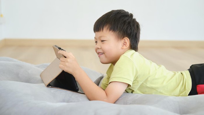 Cute little Asian kindergarten  boy child lying on his stomach while playing game, watching cartoon, using tablet pc computer, Gadget addicted children, Learning Tablet for Kids - Selective focus