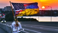 Olga Conover dari Palm Bay, Florida, berjalan menyusuri jembatan saat matahari terbenam sambil membawa bendera gabungan Amerika-Ukraina menjelang akhir pawai yang menandai peringatan satu tahun Rusia menginvasi Ukraina di Melbourne. AP/Malcolm Denemark